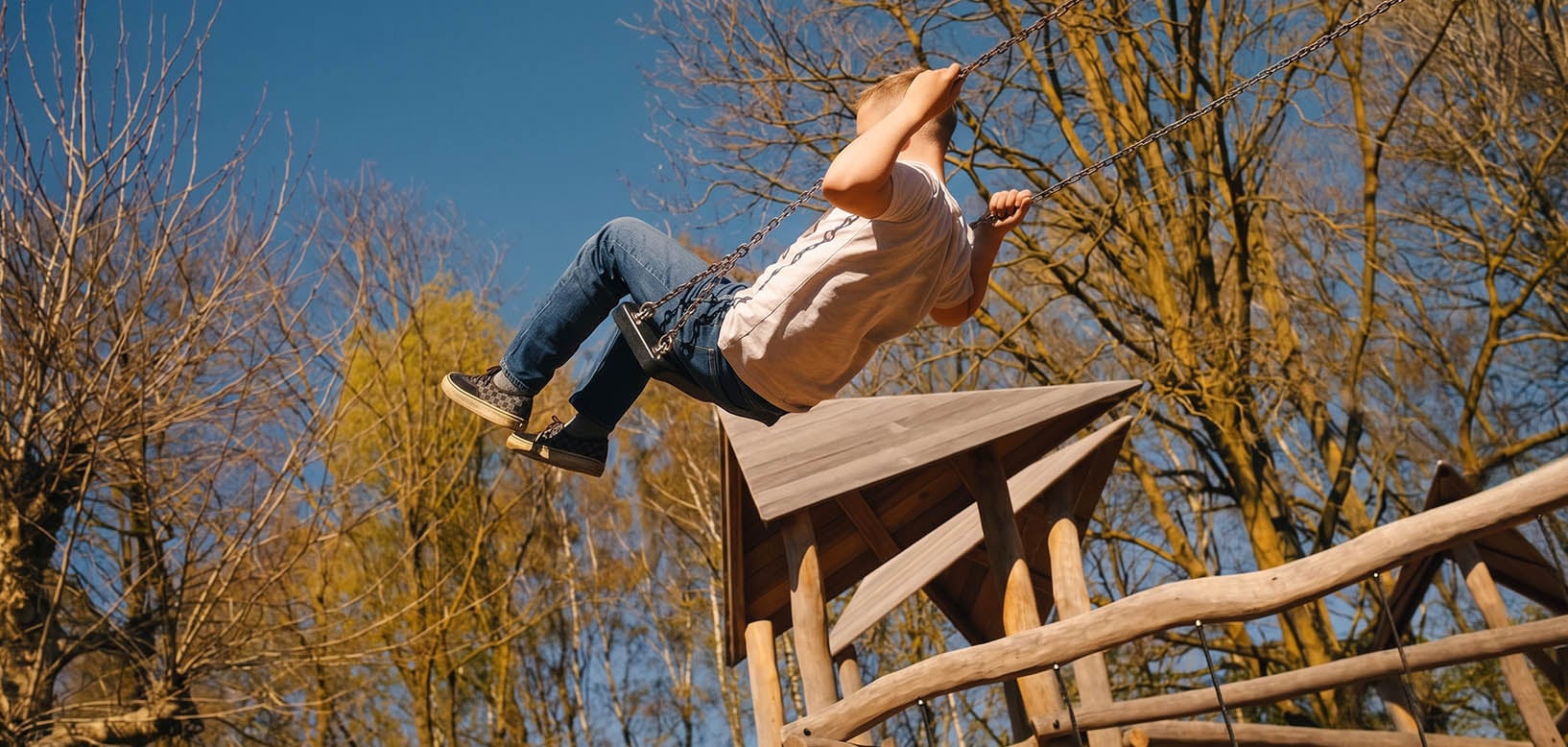 Kind schommelt hoog op een houten speeltoestel omringd door bomen in een groene speeltuin.
