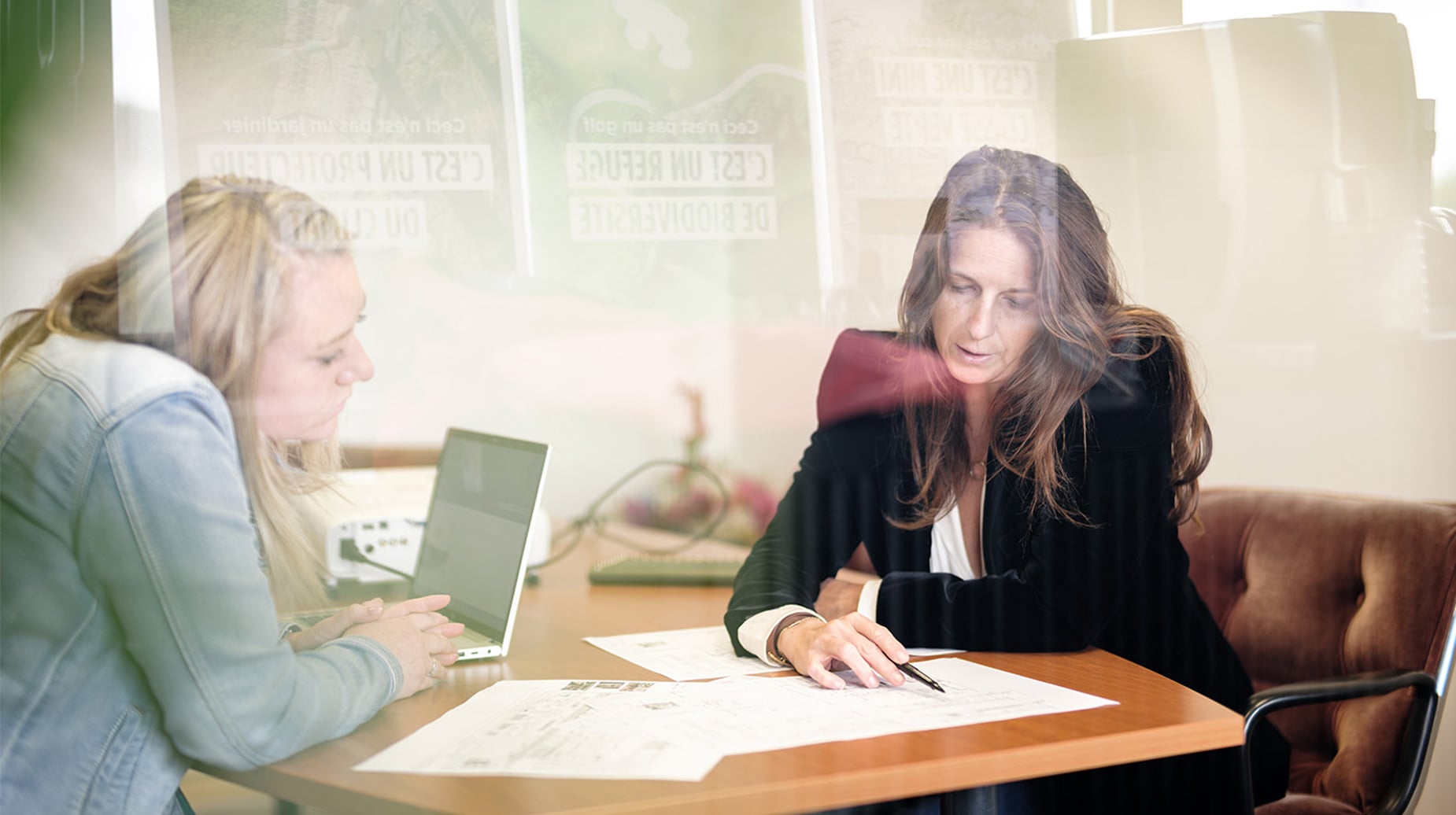 Twee medewerkers bespreken een ontwerptekening op kantoor achter glas met Franstalige groenposters.
