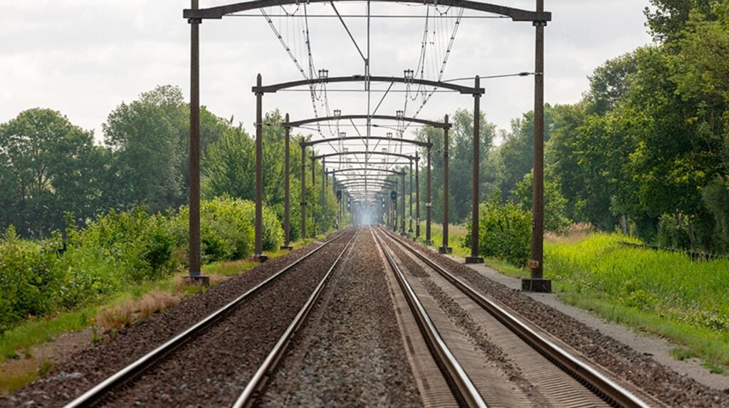 Spoorlijn met bovenleiding, geflankeerd door onderhouden groenstroken en struiken.