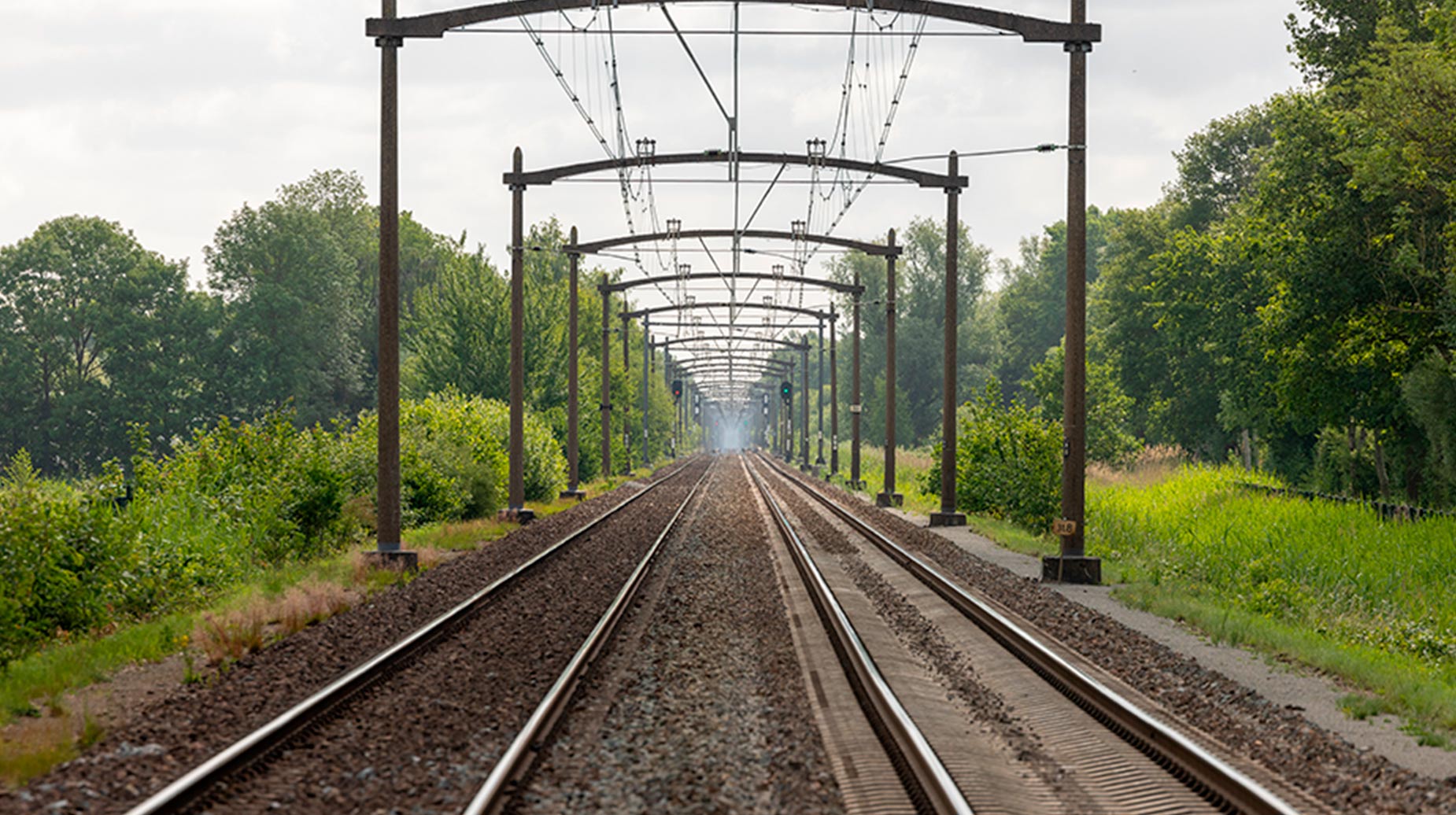 Spoorlijn met bovenleiding, geflankeerd door onderhouden groenstroken en struiken.
