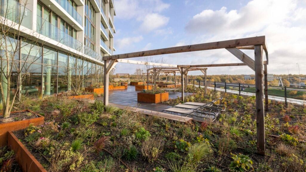 Beplant daktuin met houten pergola's en cortenstalen plantenbakken naast een hoogbouw.