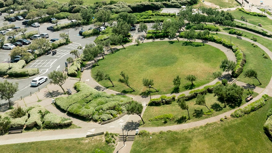 Luchtfoto van een rond grasveld omringd door bomen, hagen en wandelpaden naast een parkeerterrein.