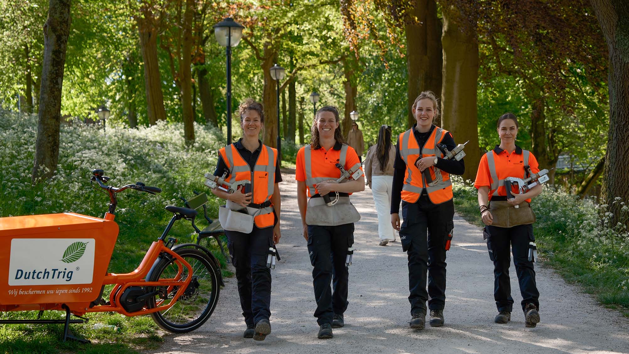 Vier medewerkers met injectieapparatuur voor iepenziektebestrijding lopen door een parklaan naast een DutchTrig-bakfiets.