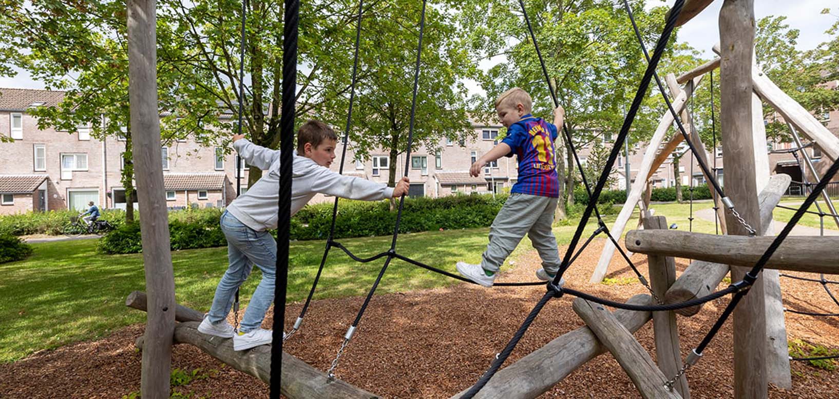 Kinderen klimmen op een houten klimtoestel met touwen in een groen wijkspeeltuintje.