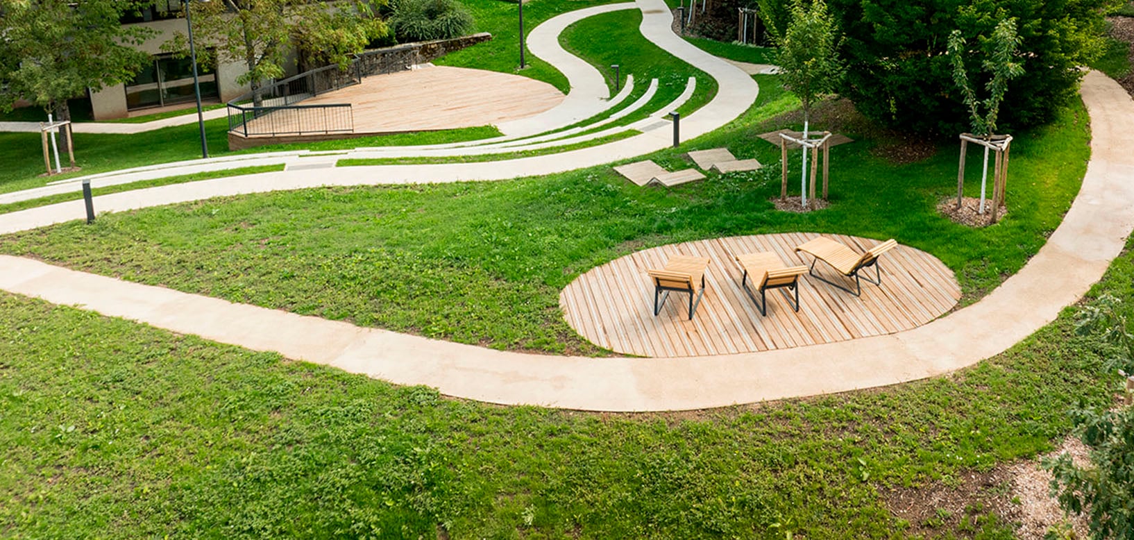 Luchtfoto van een parkaanleg met slingerende paden, gazon en een rond houten terras met ligstoelen.