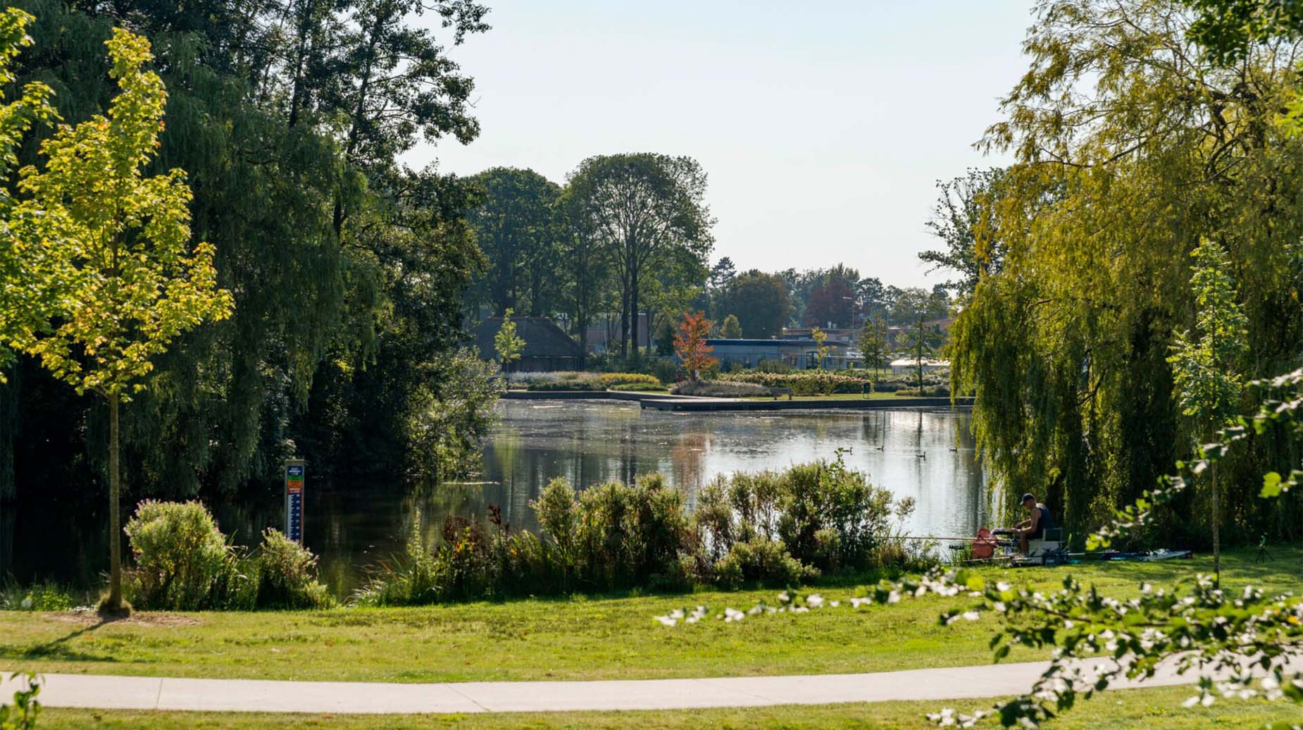 Groen ingericht park met vijver, grasveld, beplanting en een visser aan de waterkant.