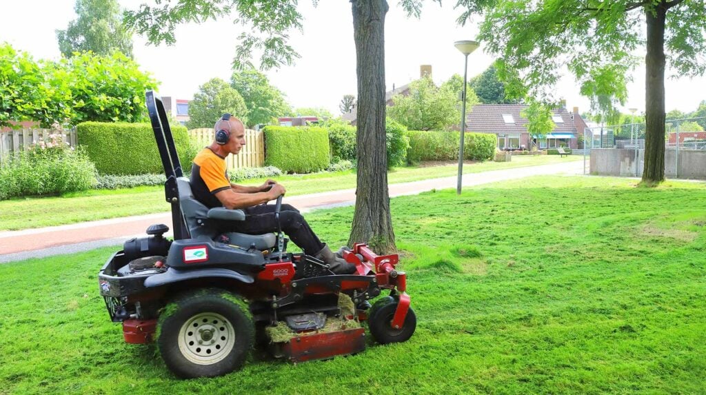 Medewerker maait gazon in woonwijk met zitmaaier tussen bomen en gazon.