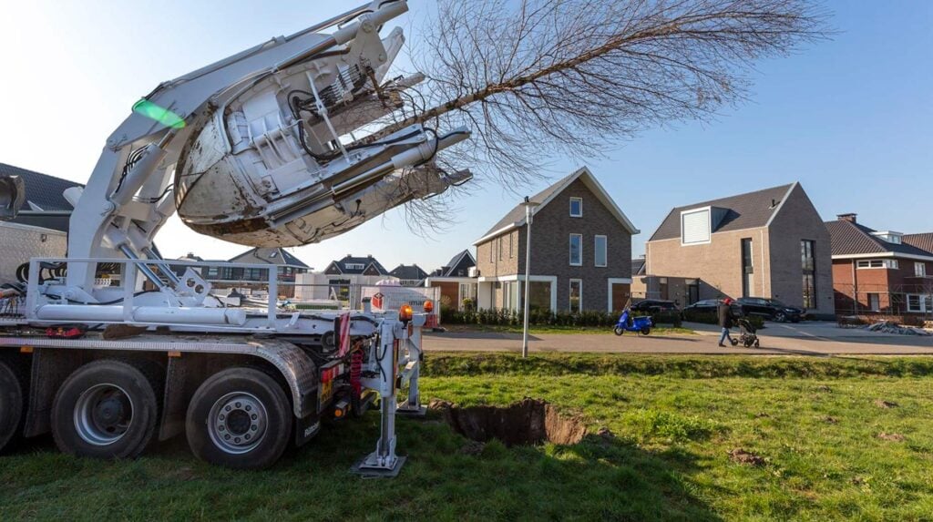Boomverplantmachine hijst een kale boom met kluit op een grasveld in een nieuwbouwwijk.