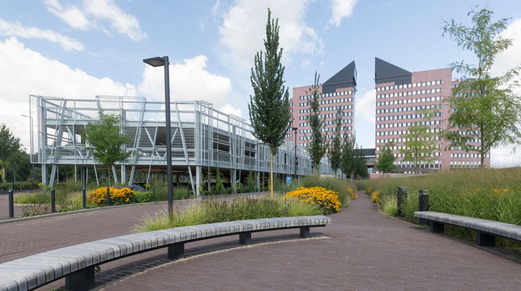 Beplant plantsoen met siergrassen en gele bloemen tussen wandelpaden bij kantoorgebouwen en een parkeergarage.