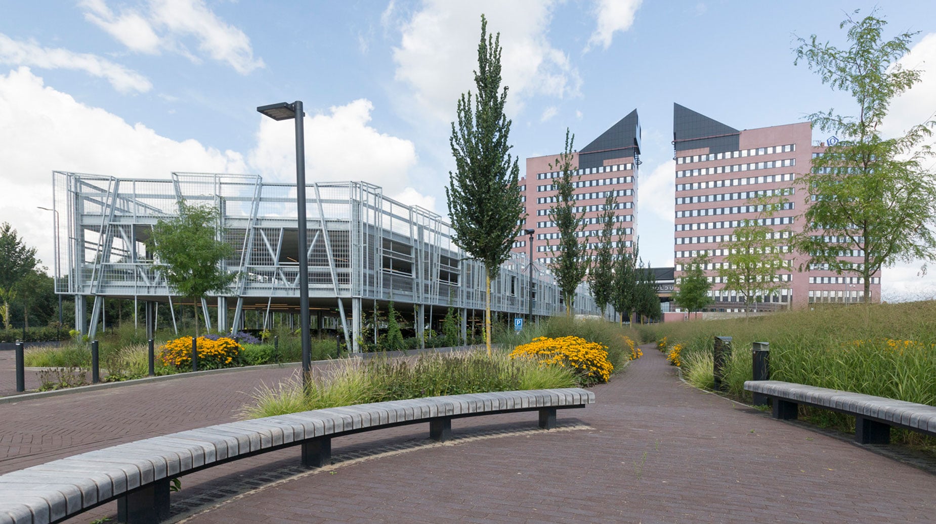 Beplant plantsoen met siergrassen en gele bloemen tussen wandelpaden bij kantoorgebouwen en een parkeergarage.