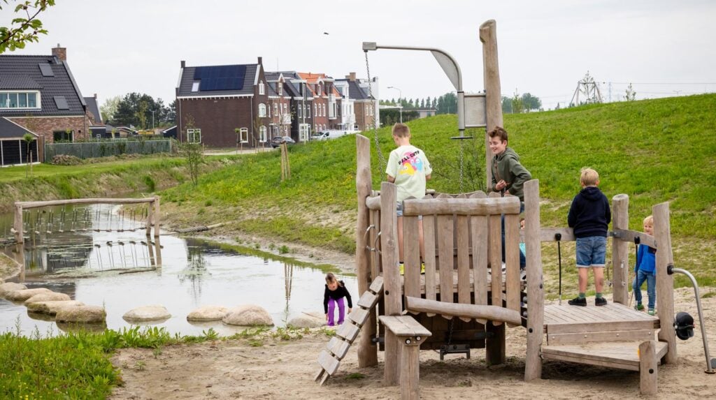 Kinderen spelen op houten speeltoestel bij waterpartij in groene woonwijk, aangelegd door idverde.