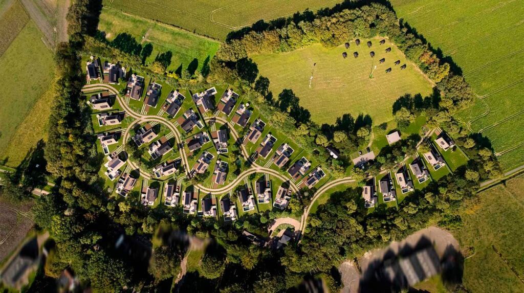 Luchtfoto van vakantiepark met bungalows in groene omgeving, omzoomd door bomen en akkers.