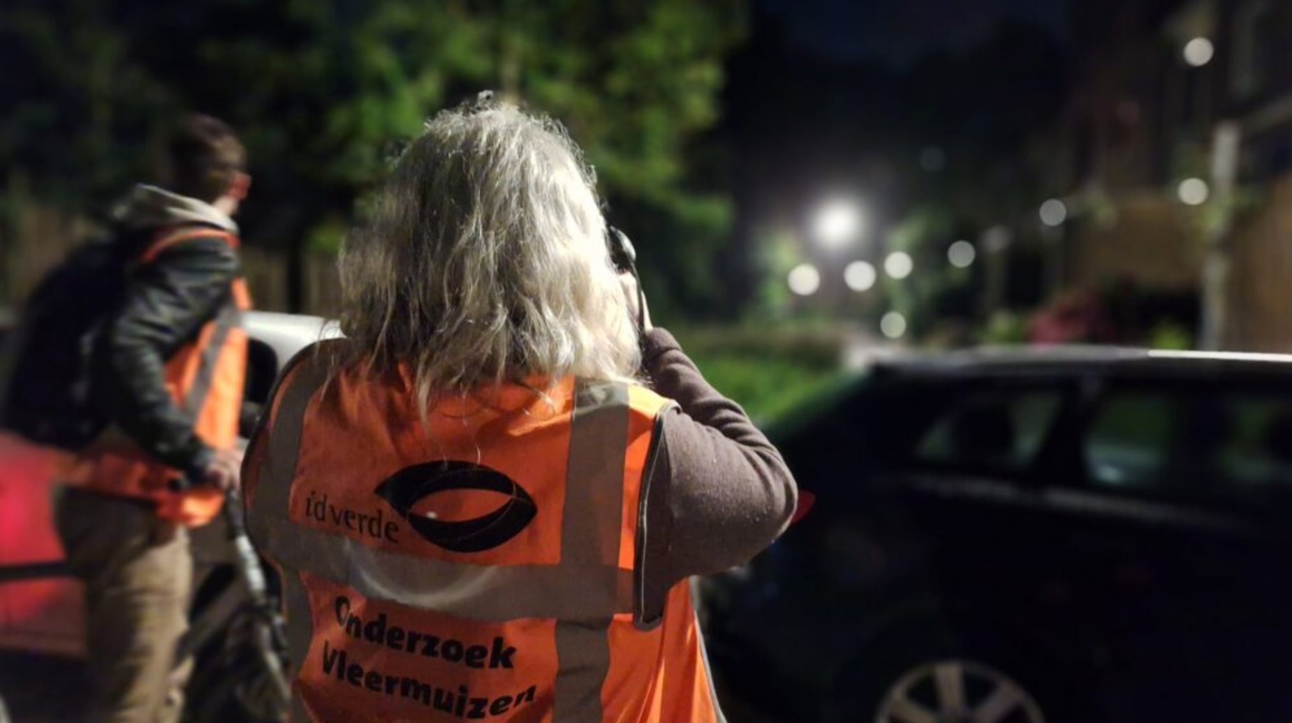 Idverde-medewerker met oranje vest 'Onderzoek Vleermuizen' luistert via koptelefoon tijdens nachtelijk veldonderzoek in Amsterdam.