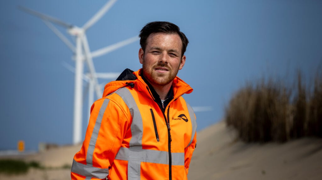 idverde-medewerker in oranje werkjas bij duinlandschap met windturbines op de achtergrond.