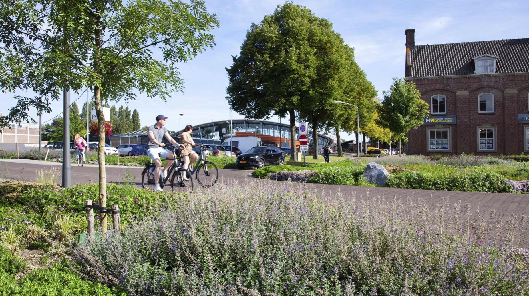 Beplant stationsplein met bloeiende border en fietsers op het fietspad langs bomen en een historisch gebouw.