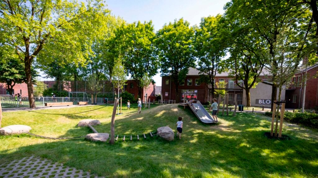 Groene speeltuin met glijbaan, klimtouw en zwerfkeien tussen bomen bij een school in de stad.