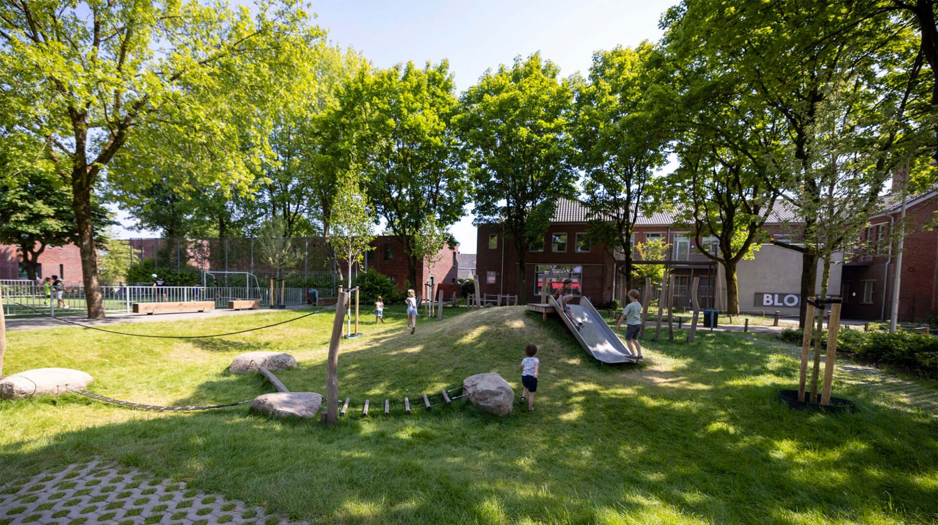 Groene speeltuin met glijbaan, klimtouw en zwerfkeien tussen bomen bij een school in de stad.