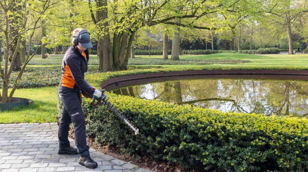 Medewerker van idverde snoeit een lage heg met een heggenschaar langs een vijver in een park.