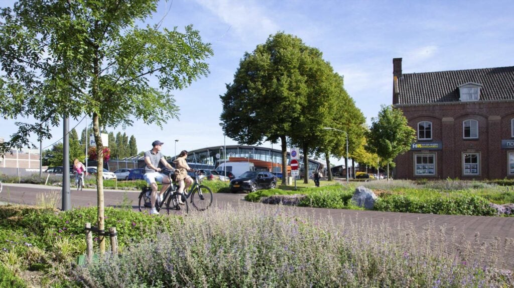 Fietsers rijden langs beplante groenstrook met vaste planten bij een stationsomgeving.