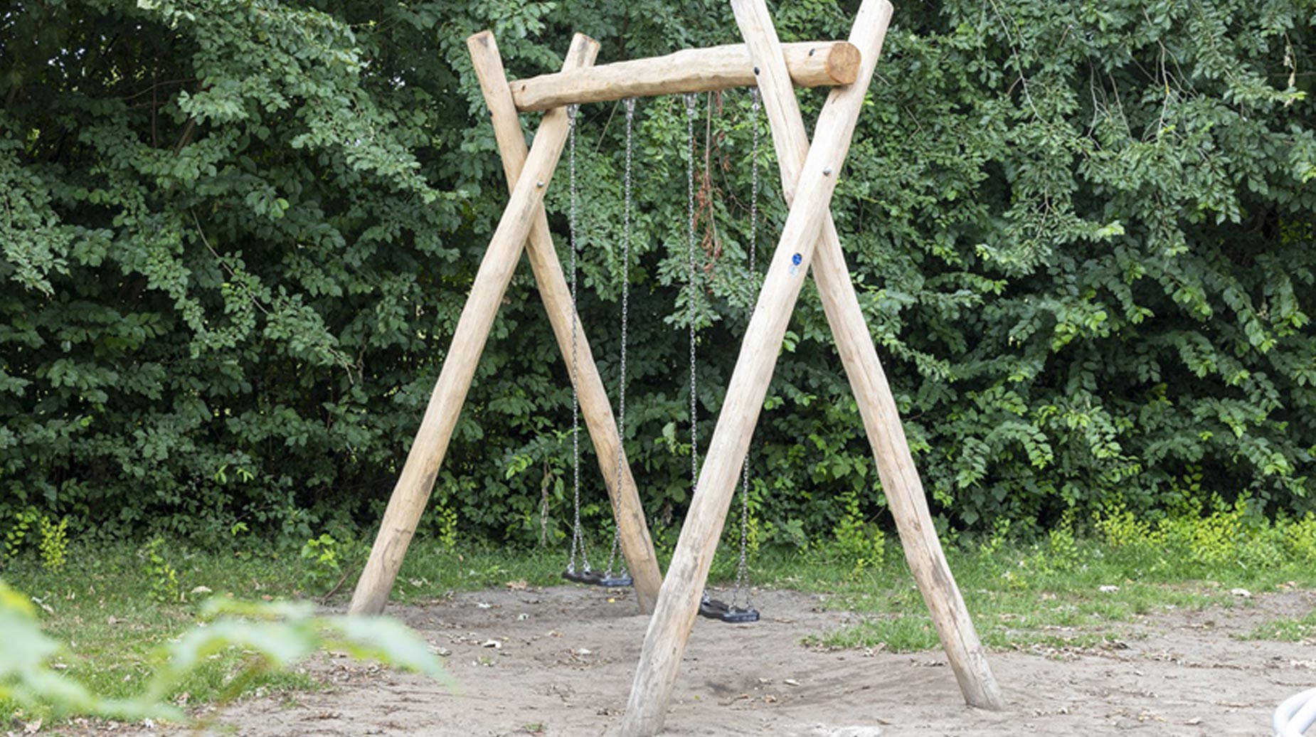 Houten schommel in openbaar groen, omringd door dichte struiken en bomen.