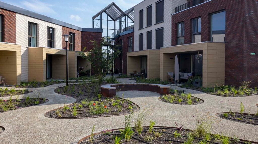 Binnentuin van een wooncomplex met slingerende grindpaden, ronde beplantingsvakken met wilde bloemen en een cortenstalen zitbank