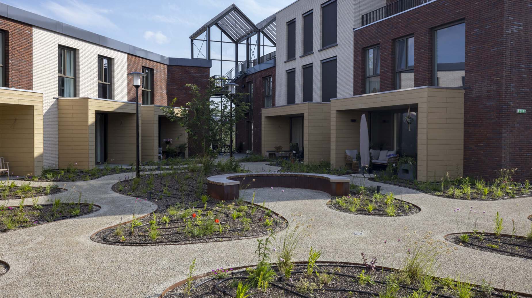 Binnentuin van een wooncomplex met slingerende grindpaden, ronde beplantingsvakken met wilde bloemen en een cortenstalen zitbank