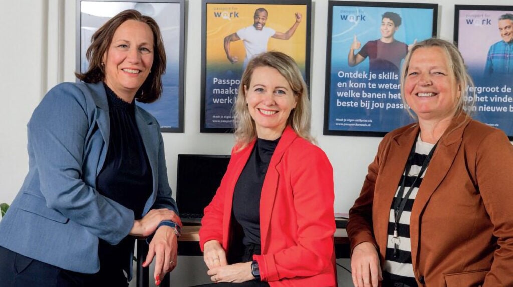Drie vrouwelijke professionals poseren glimlachend voor Passport for Work-posters in een kantooromgeving.