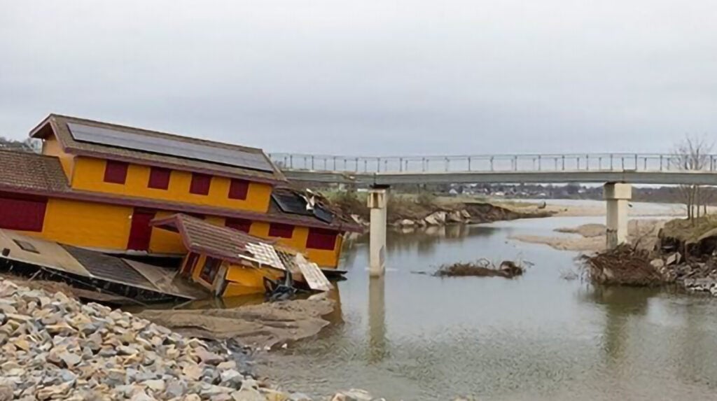 Verwoest geel houten gebouw zakt in de Maas na hoogwaterschade, met een brug op de achtergrond.