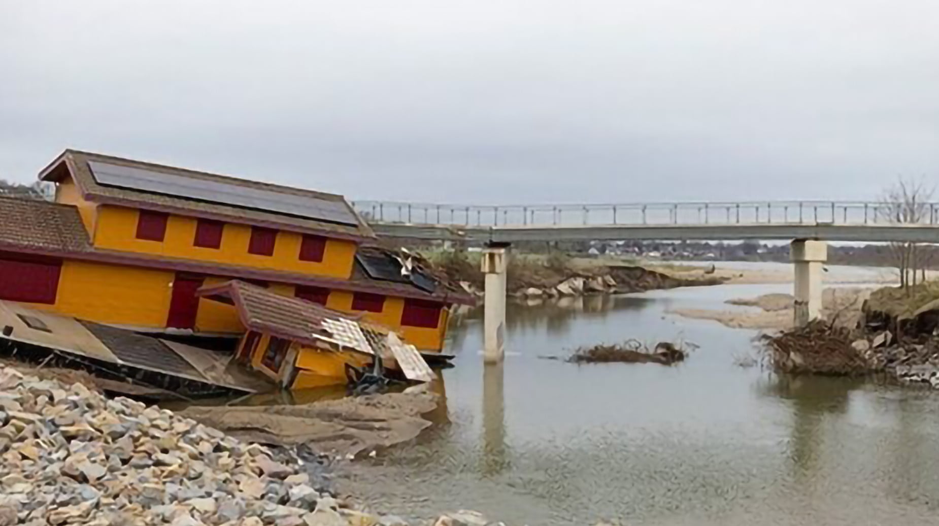 Verwoest geel houten gebouw zakt in de Maas na hoogwaterschade, met een brug op de achtergrond.