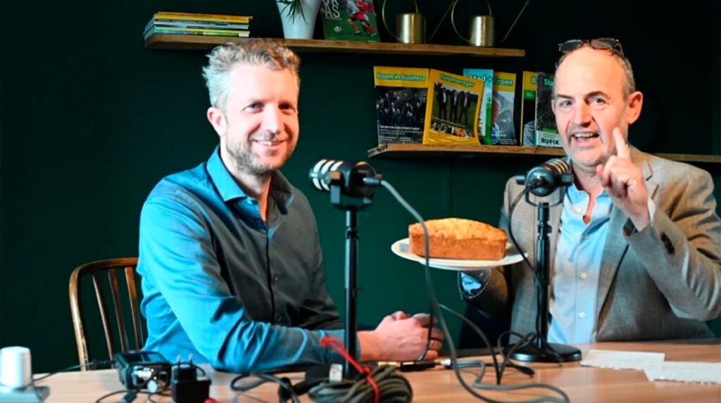 Twee mannen aan een podcasttafel met microfoons en een cake, voor een groene achtergrond met vakbladen.
