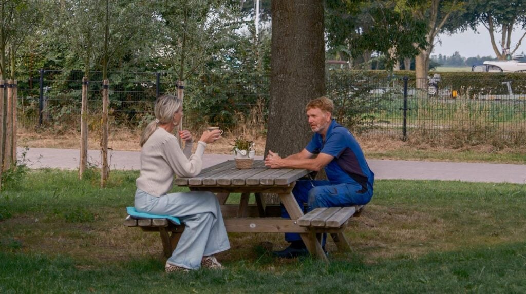 Medewerker in werkkleding overlegt met klant aan een picknicktafel bij een boom in een groene buitenruimte.
