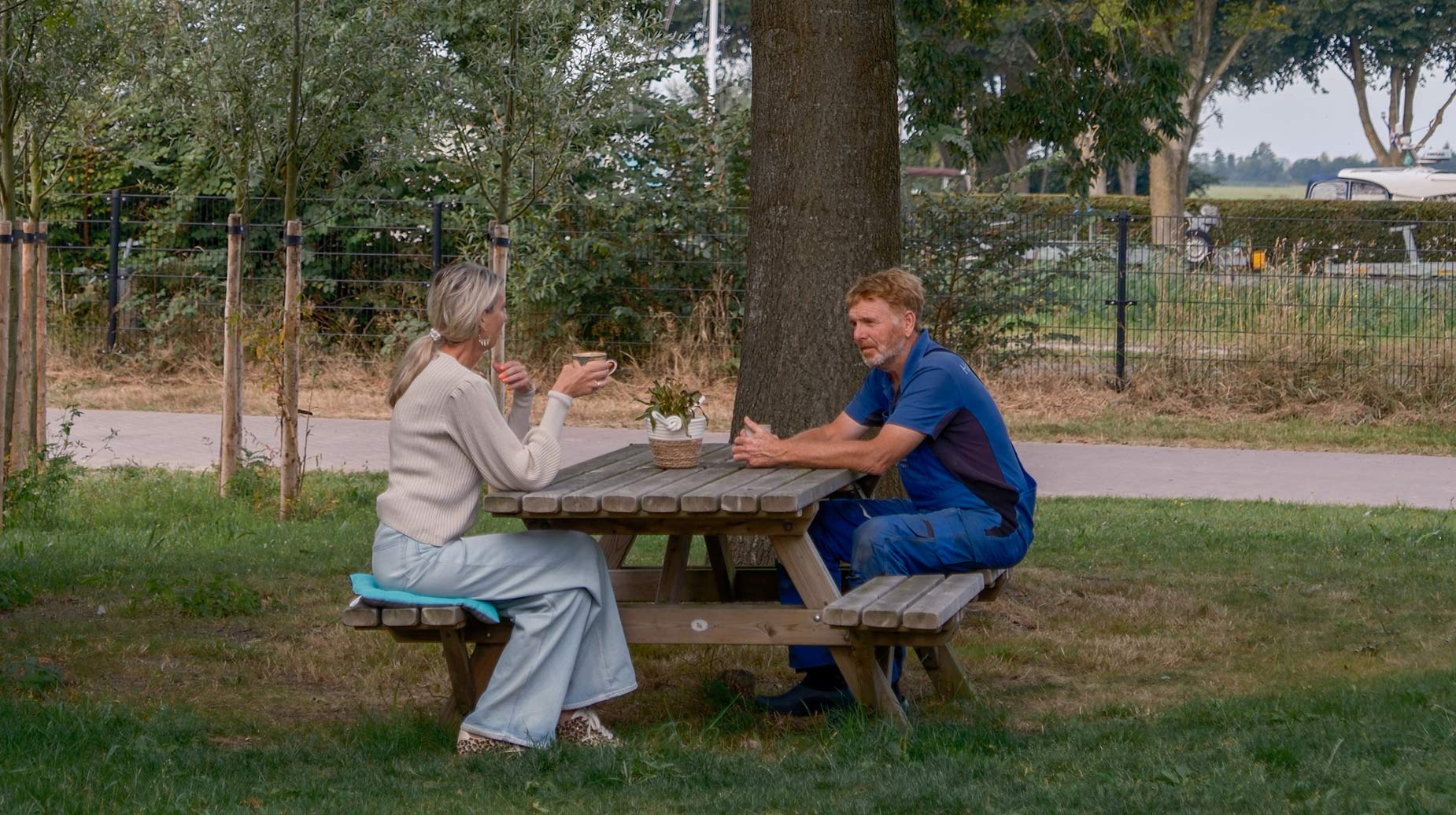 Medewerker in werkkleding overlegt met klant aan een picknicktafel bij een boom in een groene buitenruimte.